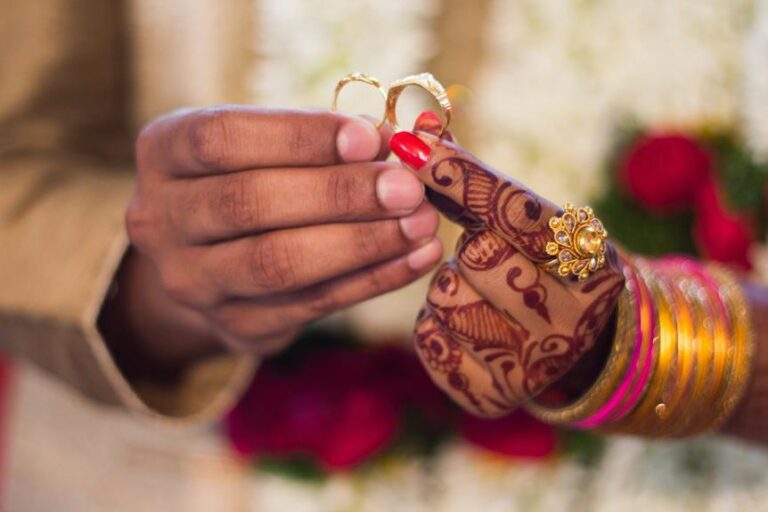 A close-up of a person's hands holding two golden rings, with intricate henna designs on her wrists and forearms, red nail polish, and several gold bangles, set against a background of flowers.