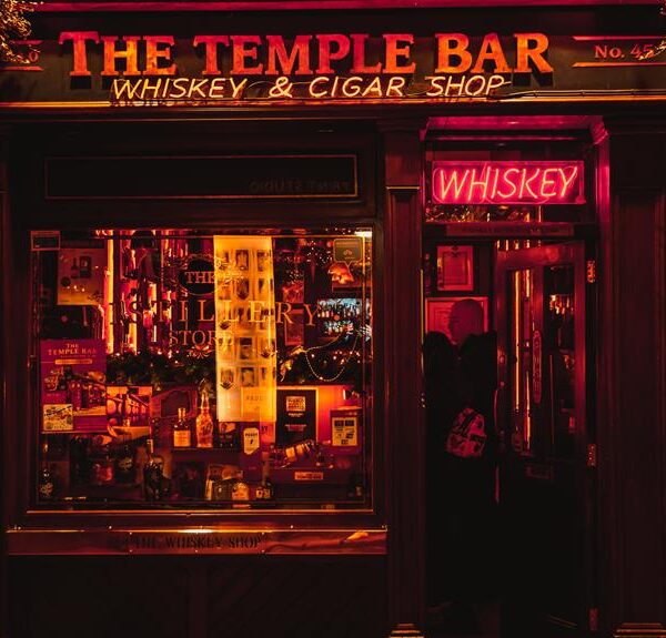 Exterior view at night of The Temple Bar Whiskey and Cigar shop with a neon whiskey sign above the door, warmly lit display window full of various bottles and goods, and dark red facade.