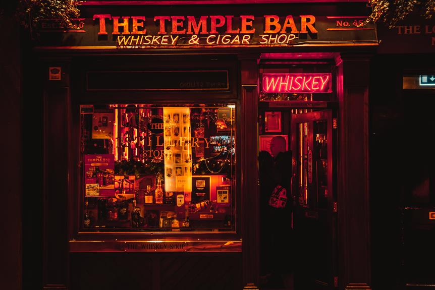 Exterior view at night of The Temple Bar Whiskey and Cigar shop with a neon whiskey sign above the door, warmly lit display window full of various bottles and goods, and dark red facade.