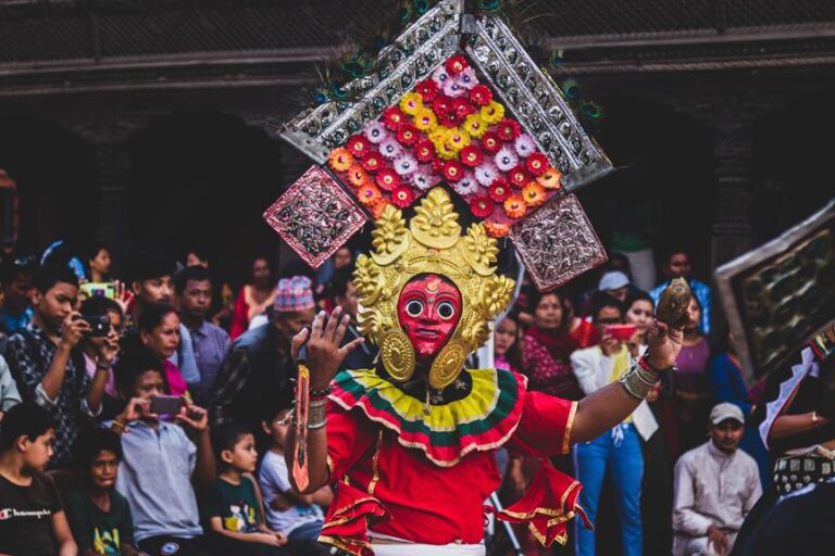 A person in traditional Nepalese costume and mask performing during a cultural festival with onlookers capturing the event on their phones.