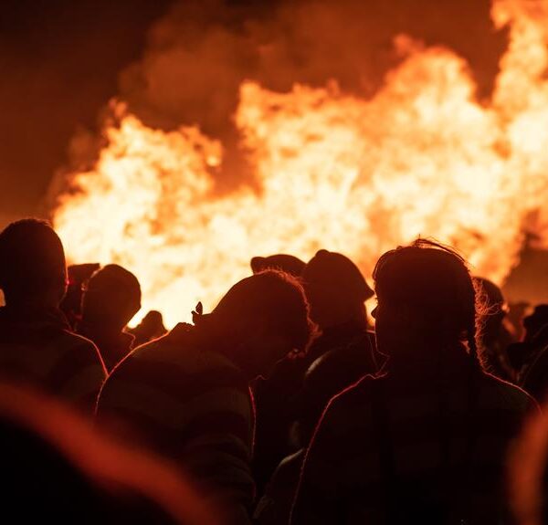Silhouetted crowd of people gathered in front of a large, intense fire at night.