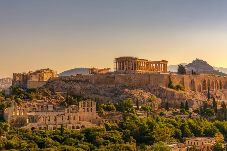 Sunset view of the Acropolis in Athens, Greece, with the Parthenon and other ancient ruins on a rocky outcrop above the city, with Lycabettus Hill in the background.